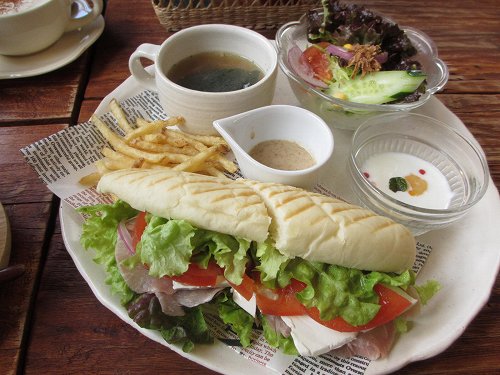 白い皿にのった細長いパンのサンド。生ハムとチーズとレタスとトマトがはさまり、スープ・サラダ・ポテトが添えられている