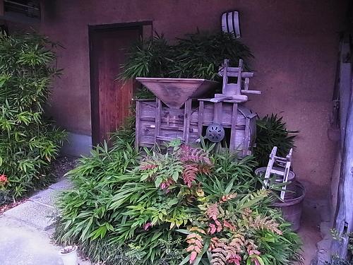 店の入口わきの植え込みに置かれた、木でできた古い唐箕（とうみ）と農具