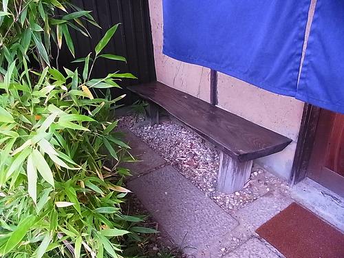 青いのれんの横に置かれた、木の長いすと笹の植え込み